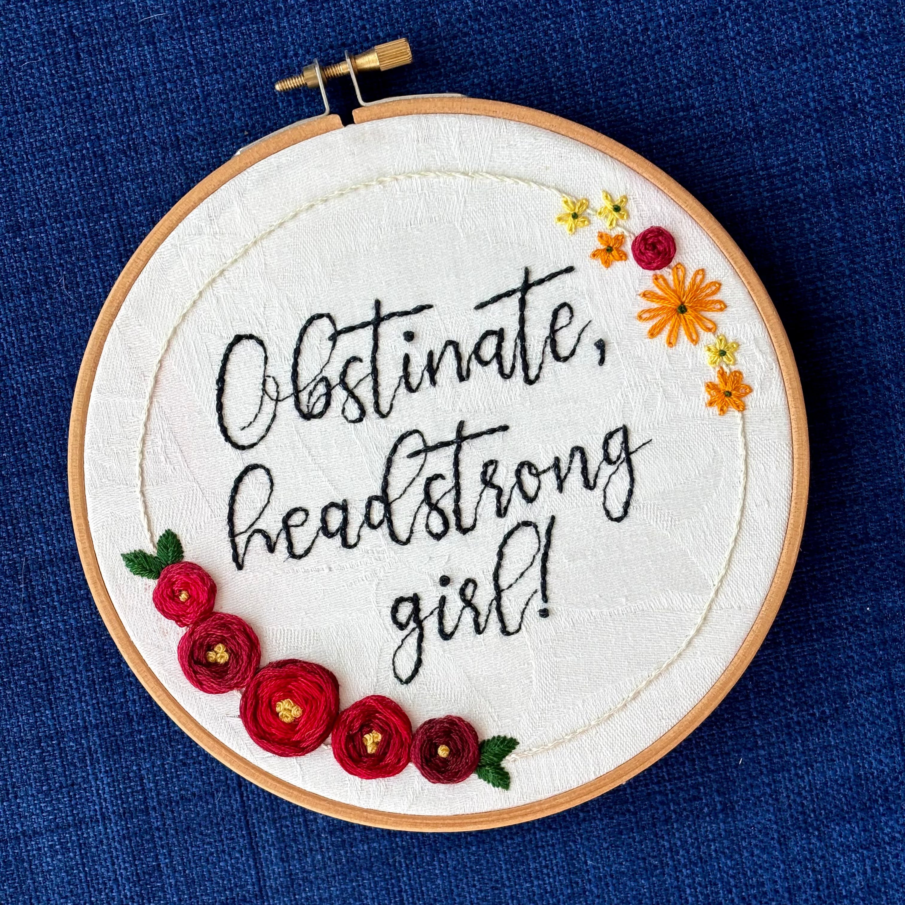 "Obstinate, headstrong girl!" Quote hand embroidered in sparkle black thread surrounded by a floral wreath with red roses and yellow and orange daisies. Fabric is white damask. Embroidery is displayed as wall art in light wood embroidery hoop.