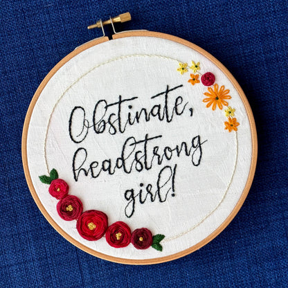 "Obstinate, headstrong girl!" Quote hand embroidered in sparkle black thread surrounded by a floral wreath with red roses and yellow and orange daisies. Fabric is white damask. Embroidery is displayed as wall art in light wood embroidery hoop.