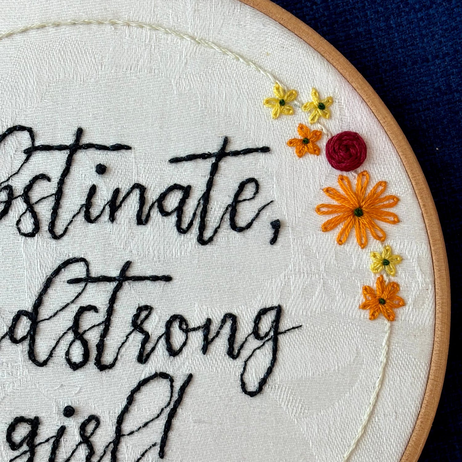 "Obstinate, headstrong girl!" Quote hand embroidered in sparkle black thread surrounded by a floral wreath with red roses and yellow and orange daisies. Fabric is white damask. Embroidery is displayed as wall art in light wood embroidery hoop.