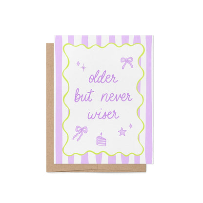 Pastel purple and white striped birthday card with the phrase "older but never wiser" in playful lavender script, featuring cute bow, star, and cake doodles, bordered by a wavy lime green frame, paired with a kraft envelope – perfect funny card for birthdays.