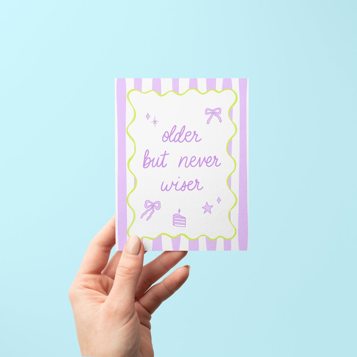 Older But Never Wiser birthday card flat lay — funny pastel birthday card styled with bow and cake doodles by indie artist MangoIllustrated