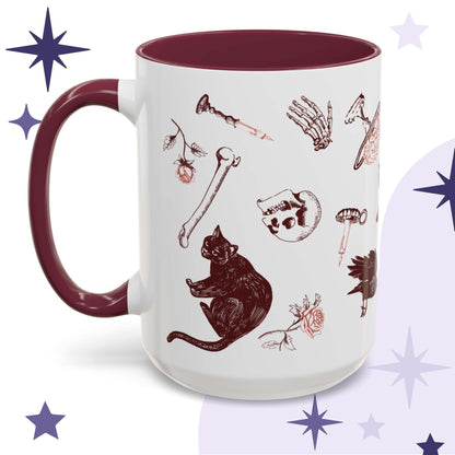 Oxblood skull and bones mug shown from the left, white ceramic scattered with red gothic motifs of black cats, ravens, skulls, and loose bones, deep burgundy interior and handle