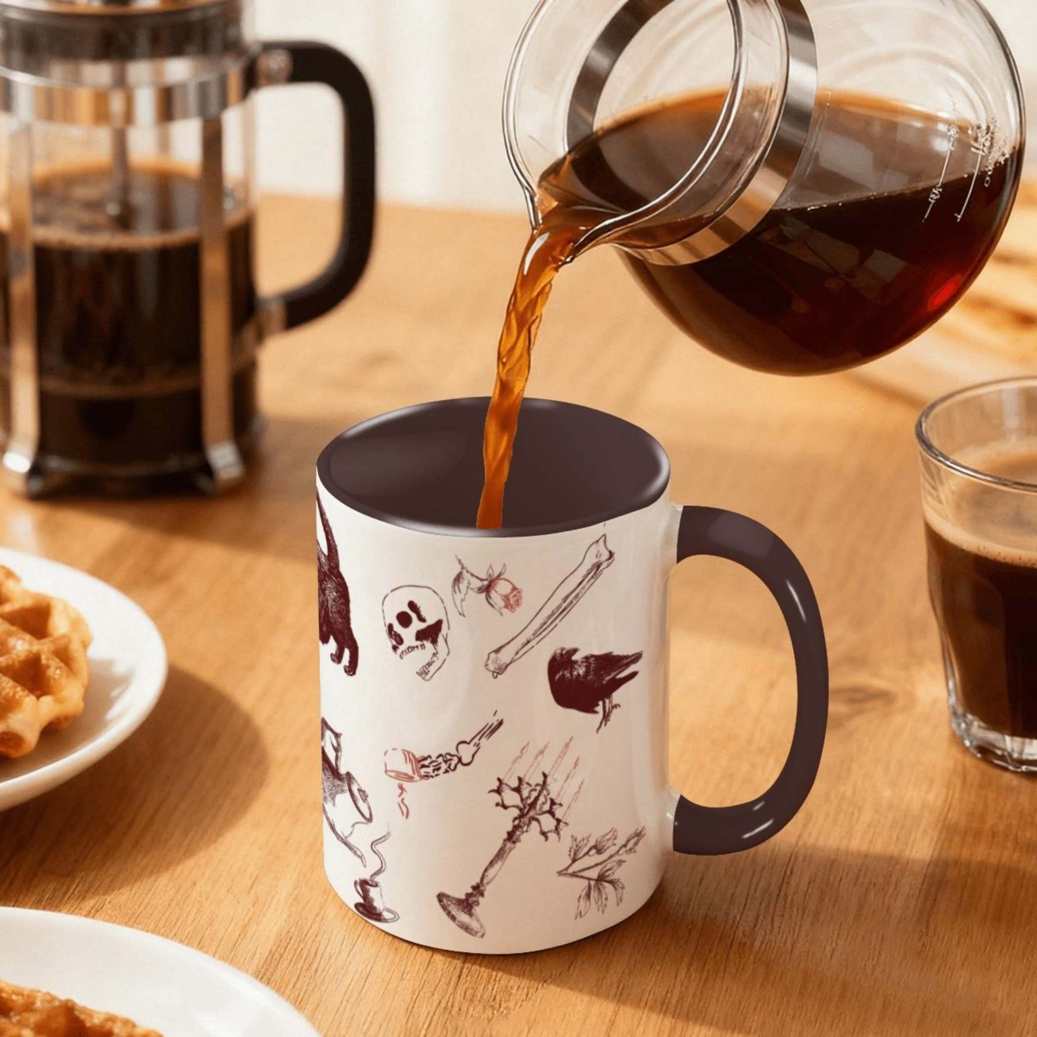 Coffee pouring into the oxblood zombie icons mug on a wood table beside a French press, red macabre scatter print of skulls, bones, and ravens
