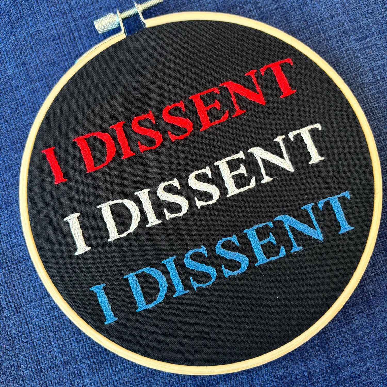Embroidery design reads, "I dissent I dissent I dissent"  Hand embroidered red, white, and blue. "I dissent" in all capital letters. Fabric is black cotton. Completed Embroidery is displayed as wall art in light wood embroidery hoop