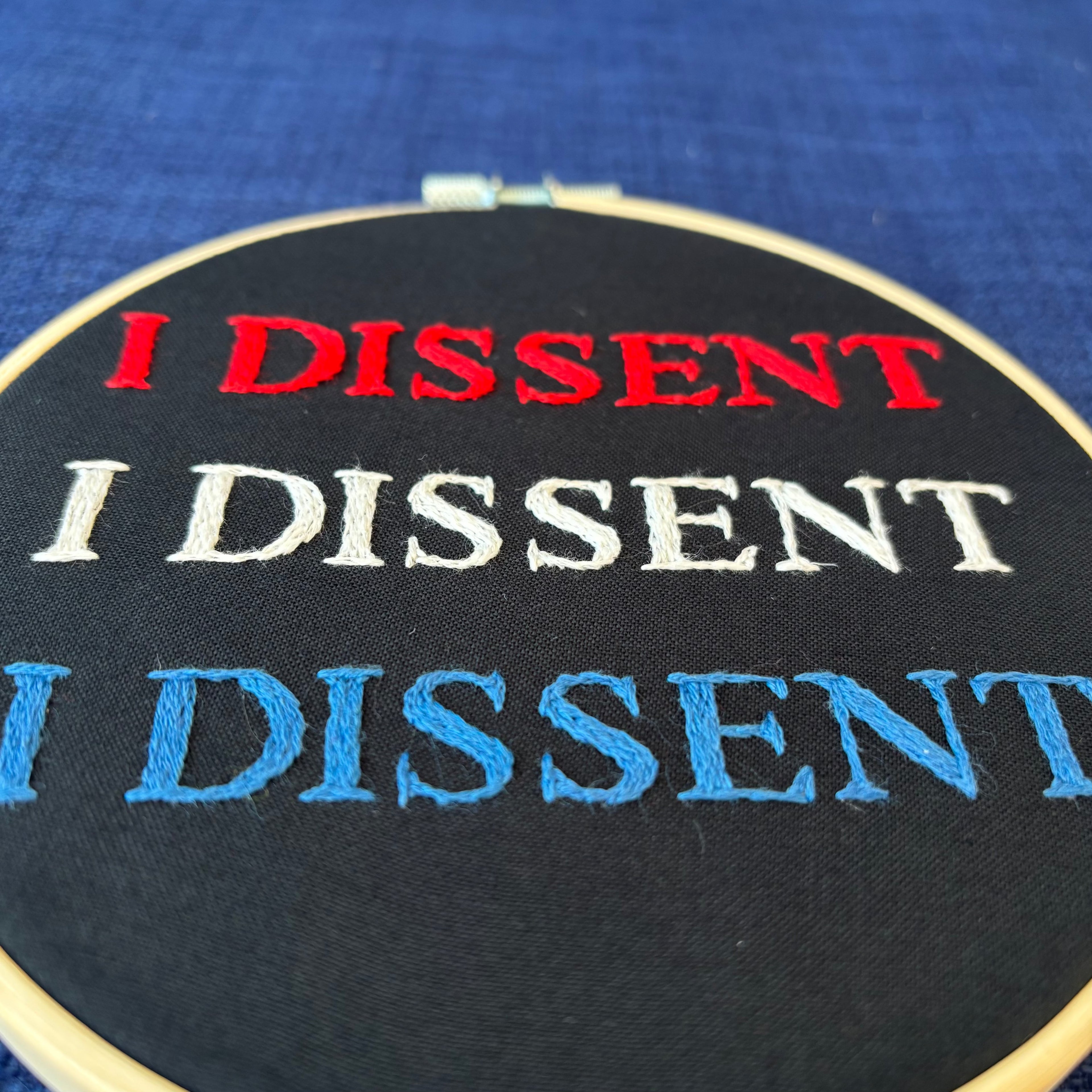 hand embroidery design reads, "I dissent I dissent I dissent"  Hand embroidered red, white, and blue. "I dissent" in all capital letters. Fabric is black cotton. Completed Embroidery is displayed as wall art in light wood embroidery hoop