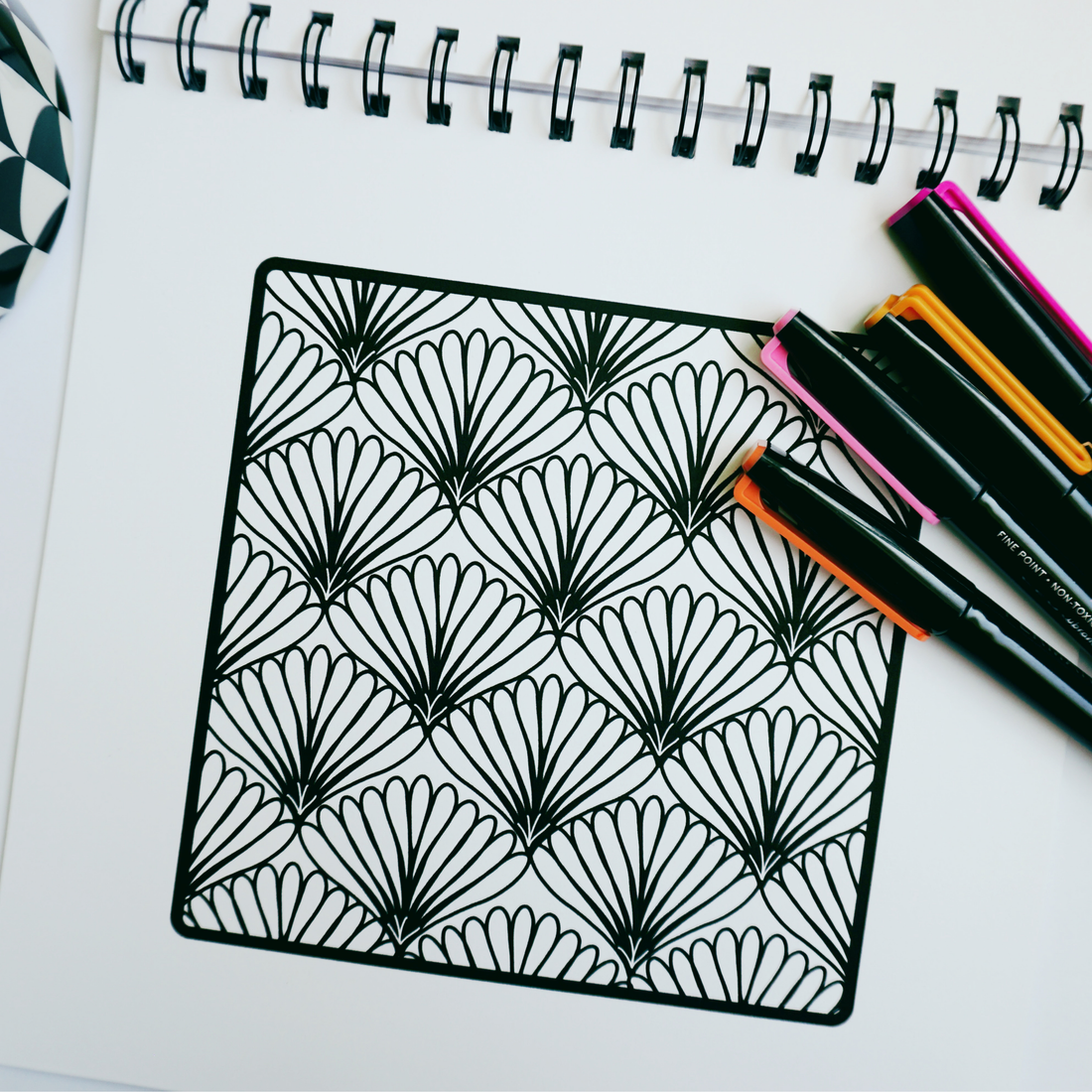 Black-and-white fan pattern drawn inside a square on a spiral coloring book page, with pink, orange, and black markers resting in the upper right corner.