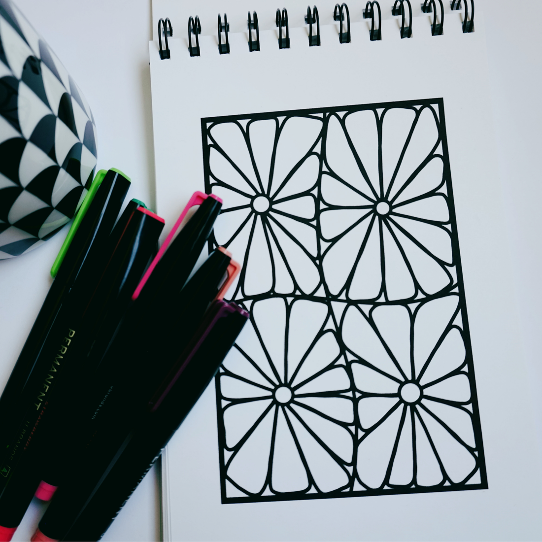 Close-up of a spiral-bound coloring book opened to a bold geometric floral design, with colorful markers and a black-and-white patterned mug beside it on a white surface. 