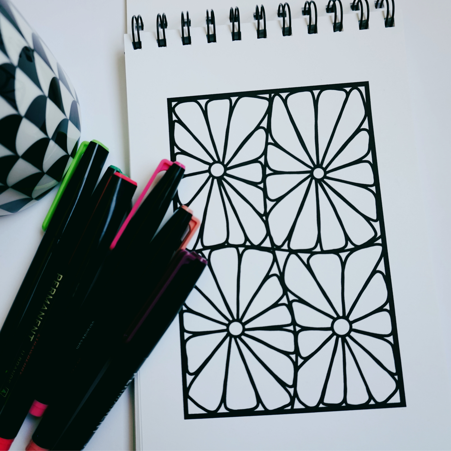 Close-up of a spiral-bound coloring book opened to a bold geometric floral design, with colorful markers and a black-and-white patterned mug beside it on a white surface. 