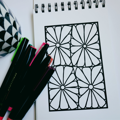 Close-up of a spiral-bound coloring book opened to a bold geometric floral design, with colorful markers and a black-and-white patterned mug beside it on a white surface. 