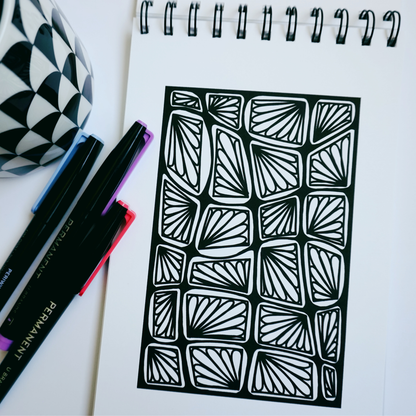 Close-up of a spiral-bound coloring book opened to a bold abstract pattern of fan-shaped designs in rounded rectangles, with colorful markers and a black-and-white patterned mug nearby.