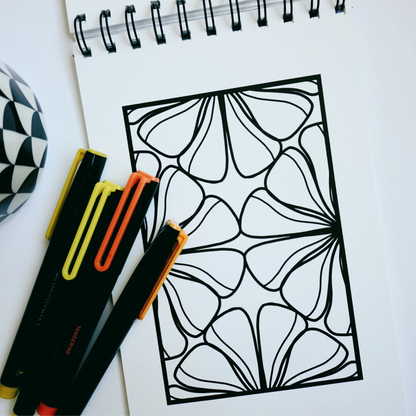 Close-up of a spiral-bound coloring book opened to an organic floral pattern with bold black outlines, alongside yellow and orange markers and a black-and-white patterned mug on a white surface.