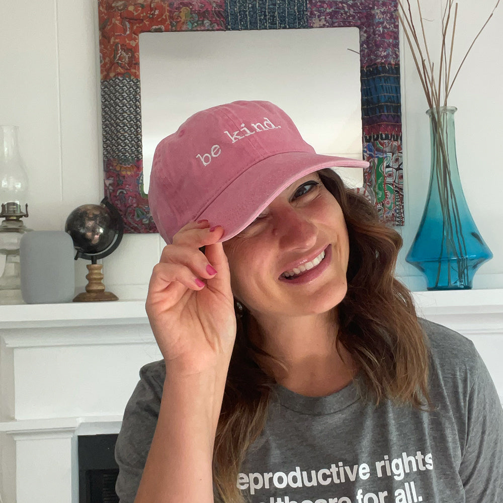 Soft pink cotton dad hat with white "be kind" embroidery, worn casually indoors by a smiling woman