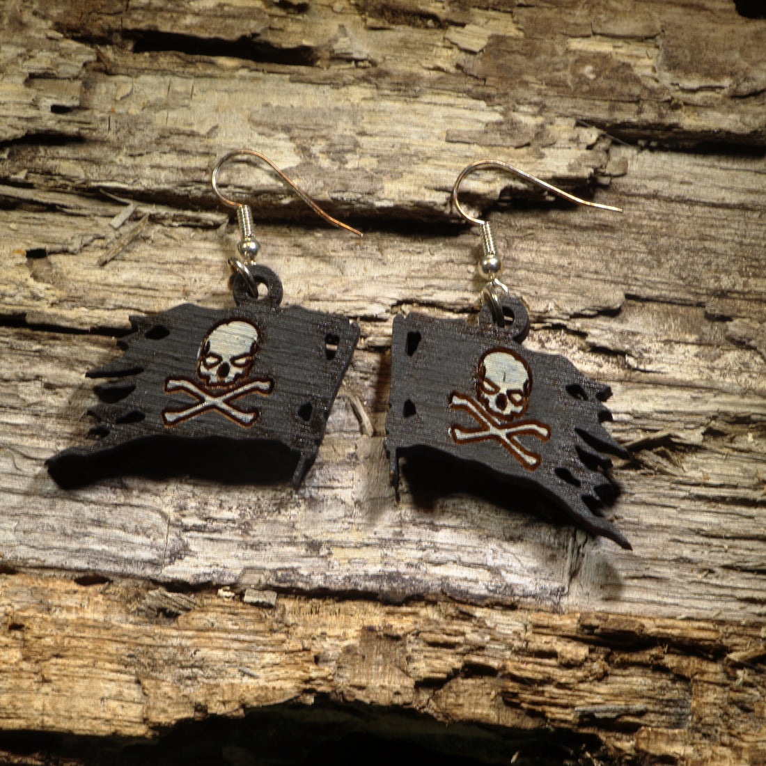 Hand-painted wooden pirate flag dangle earrings featuring the classic skull and crossbones on a weathered black flag. Crafted from lightweight wood with hypoallergenic .925 sterling silver hooks, these swashbuckling earrings are perfect for pirate enthusiasts, Renaissance festival fans, Halloween costumes, nautical lovers, and anyone who enjoys adventurous statement jewelry with a touch of high-seas flair.