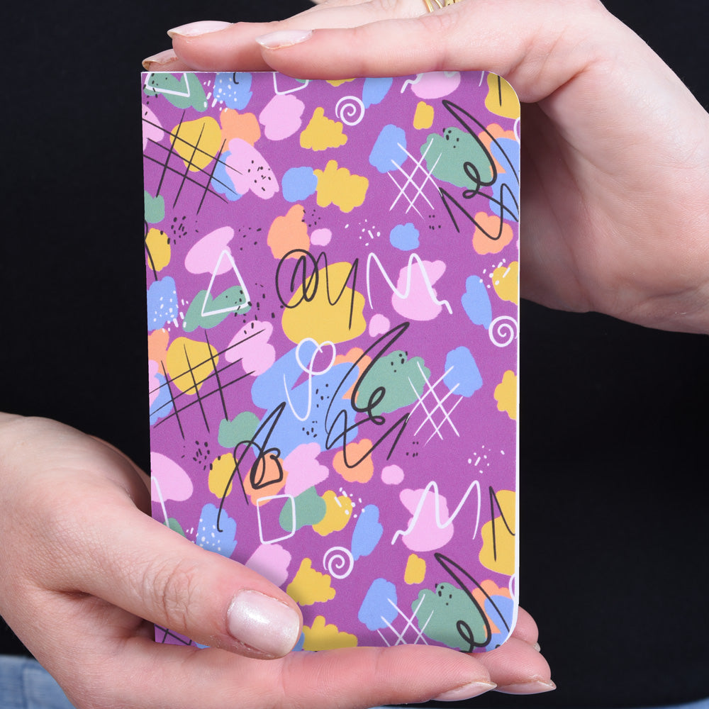 hands holding a small pocket notebook featuring a magenta abstract design with doodles and paint splotches