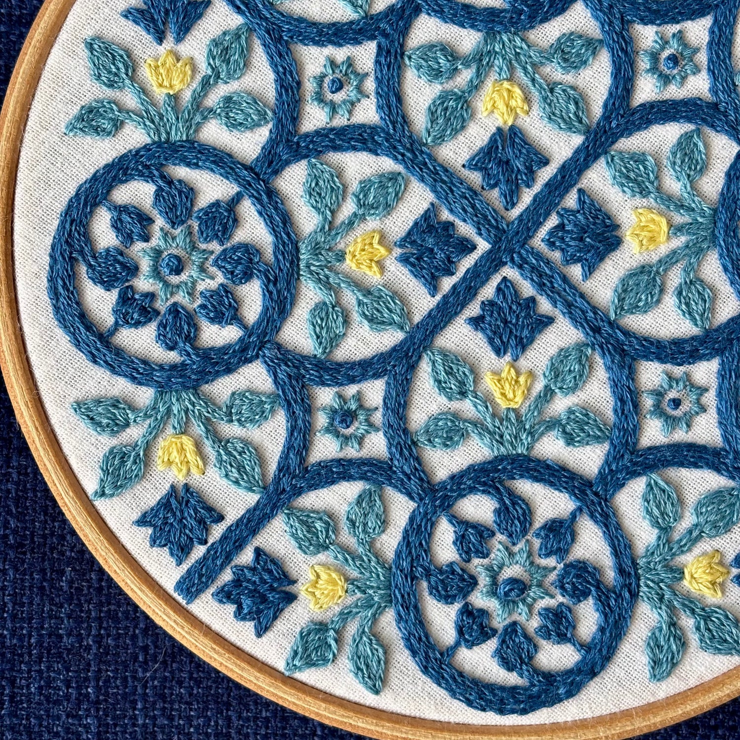 Dark blue, light blue, and yellow embroidery based on traditional Portuguese tile with flowers, leaves, lines, and stars. Fabric is white on white cotton. Embroidery is displayed as wall art in light wood embroidery hoop.