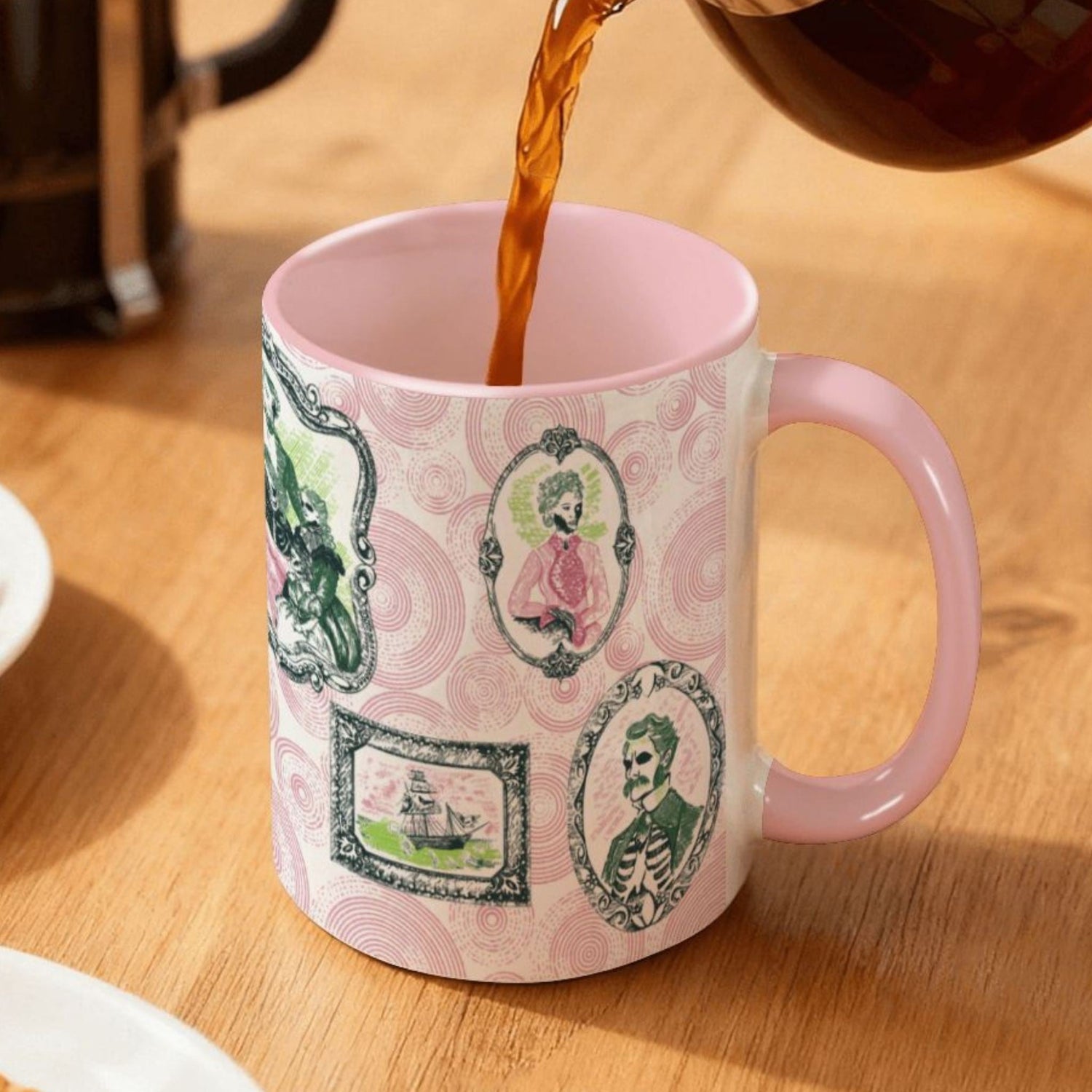 Coffee pouring into a pink-handled preppy portrait mug on a wood table, pink and green skull-faced gallery frames