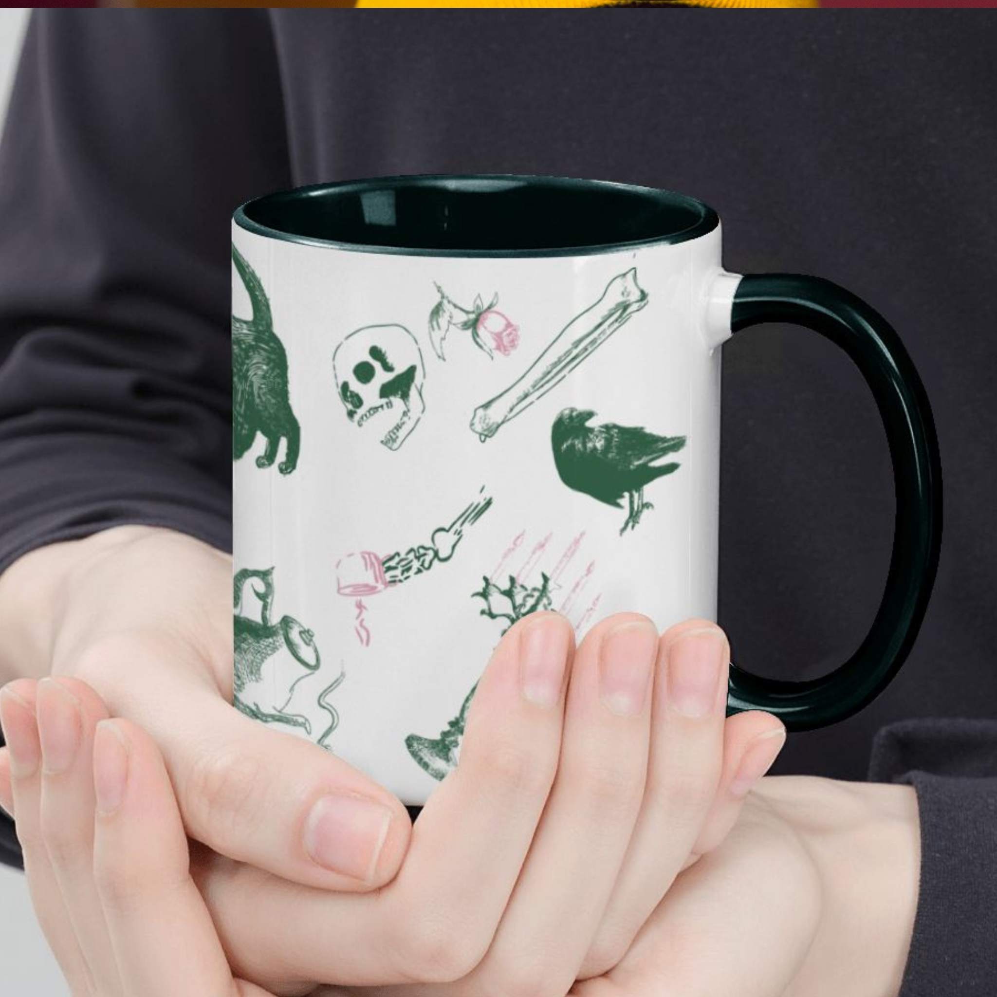 Hands cradling a forest green preppy skull and bones mug, close view of green and pink motifs including a raven, a skull, and a skeletal hand