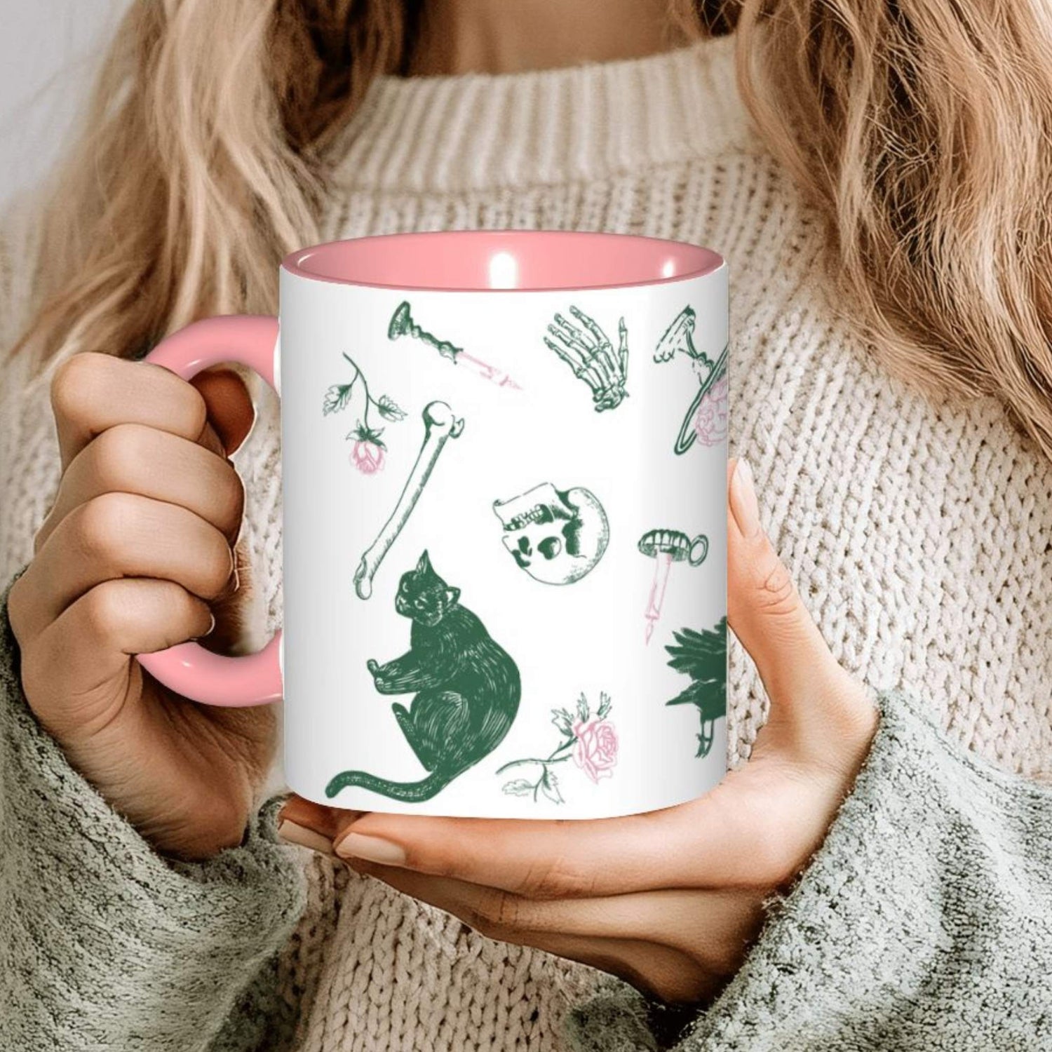 Hands holding a pink preppy skull and bones mug in a cozy sweater, green and pink motifs including a black cat, a skull, and roses