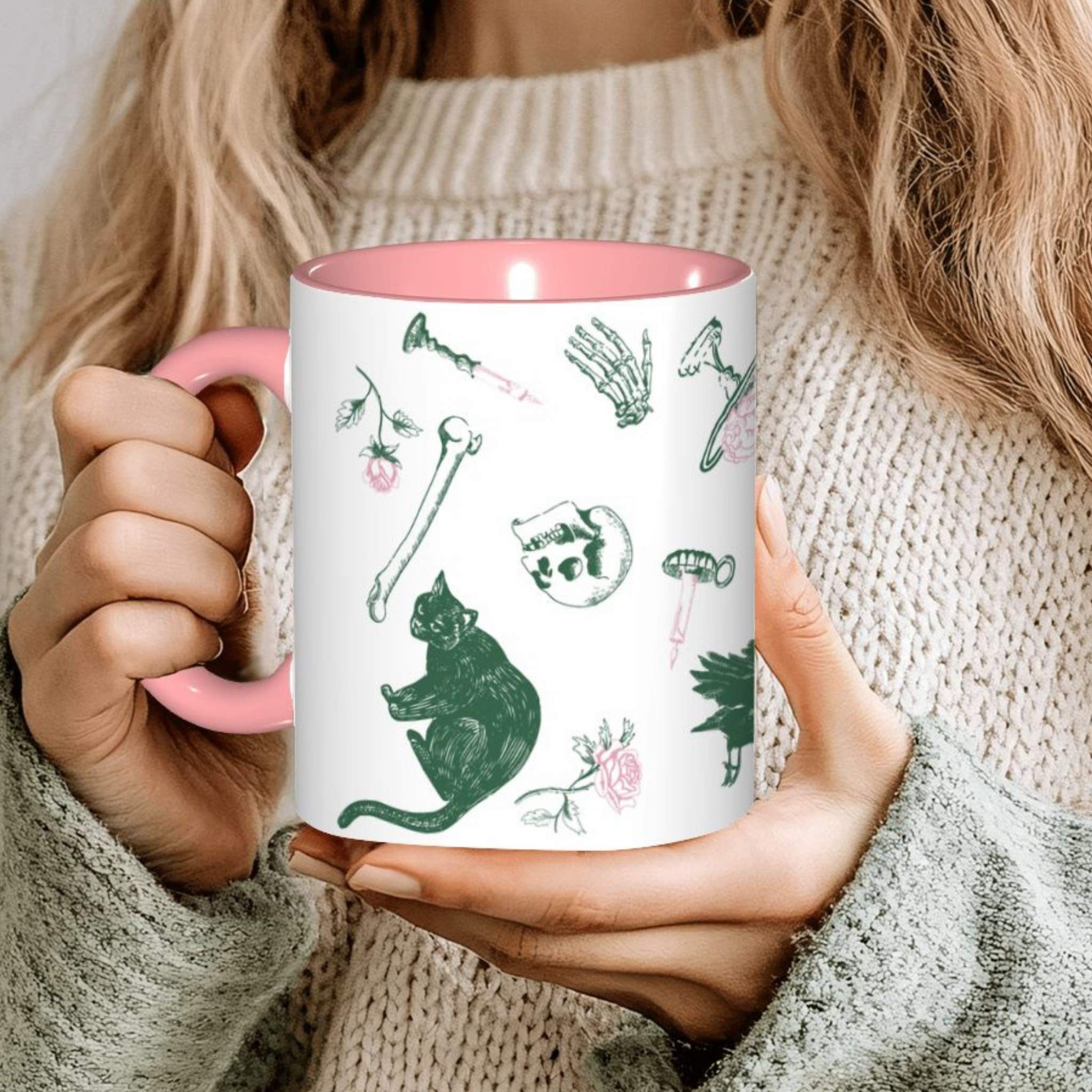 Hands holding a pink preppy skull and bones mug in a cozy sweater, green and pink motifs including a black cat, a skull, and roses