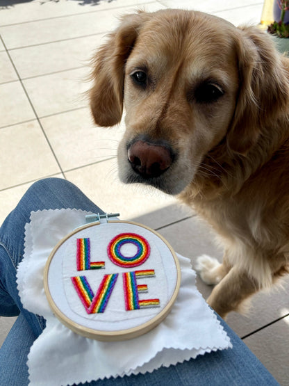 Photo of this embroidery project in my lap with my golden retriever dog looking on. The word "love" is hand embroidered on two lines, in a square, in all capital letters. Each letter is stitched in stripes of rainbow colors: pink, red, orange, yellow, green, blue, purple. Text is sewn on white cotton. Framed as wall art in light wood embroidery hoop.