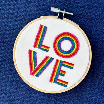 The word "love" is hand embroidered on two lines, in a square, in all capital letters. Each letter is stitched in stripes of rainbow colors: pink, red, orange, yellow, green, blue, purple. Text is sewn on white cotton. Framed as wall art in light wood embroidery hoop.