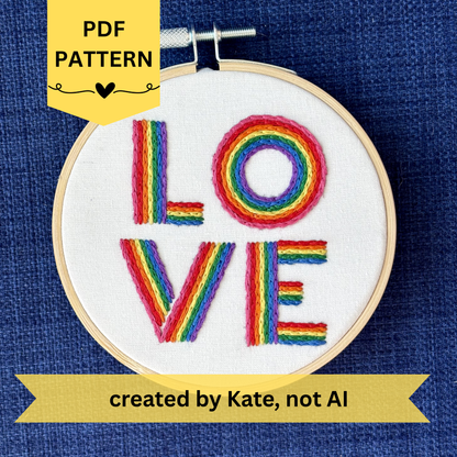 PDF pattern created by Kate, not AI. The word "love" is hand embroidered on two lines, in a square, in all capital letters. Each letter is stitched in stripes of rainbow colors: pink, red, orange, yellow, green, blue, purple. Text is sewn on white cotton. Framed as wall art in light wood embroidery hoop.