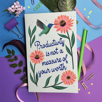 Notebook with lettering "productivity is not a measure of your worth" with a hummingbird and flowers surrounded by stationery items on a colorful background