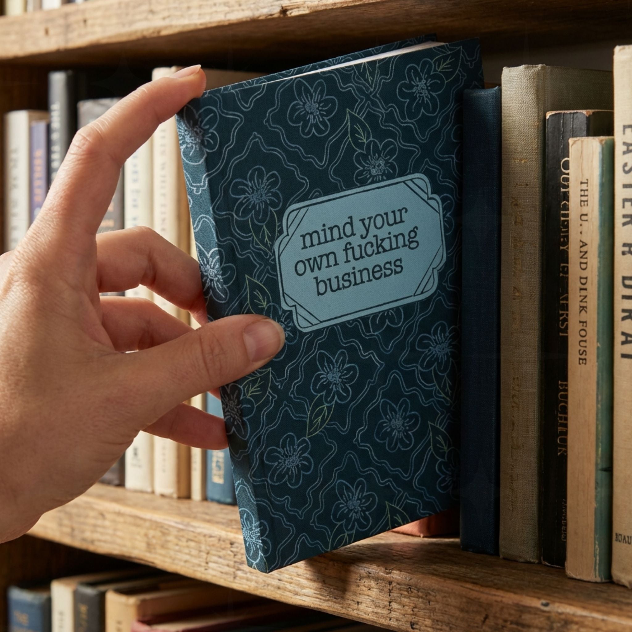 Hand pulling profanity floral journal from wooden bookshelf among vintage books showing teal botanical cover