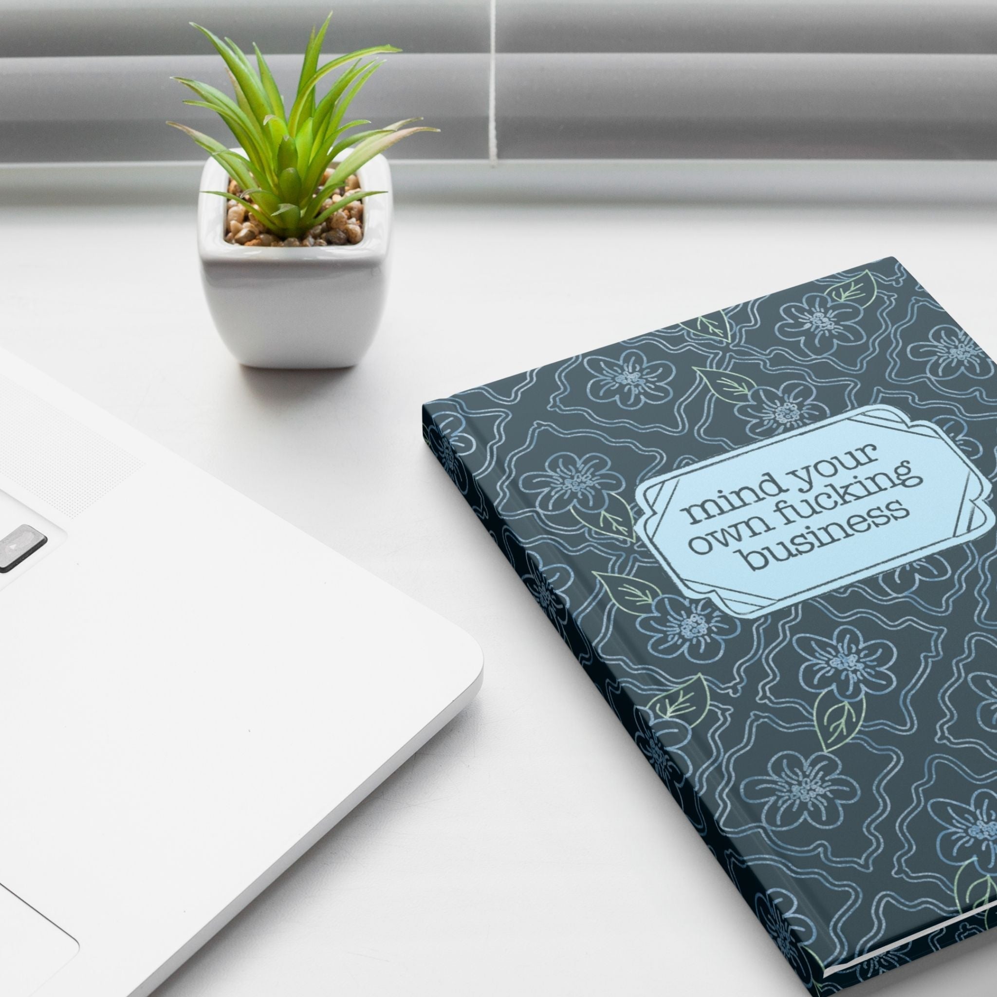 Profanity floral journal on white desk next to laptop and small succulent plant showing teal botanical hardcover