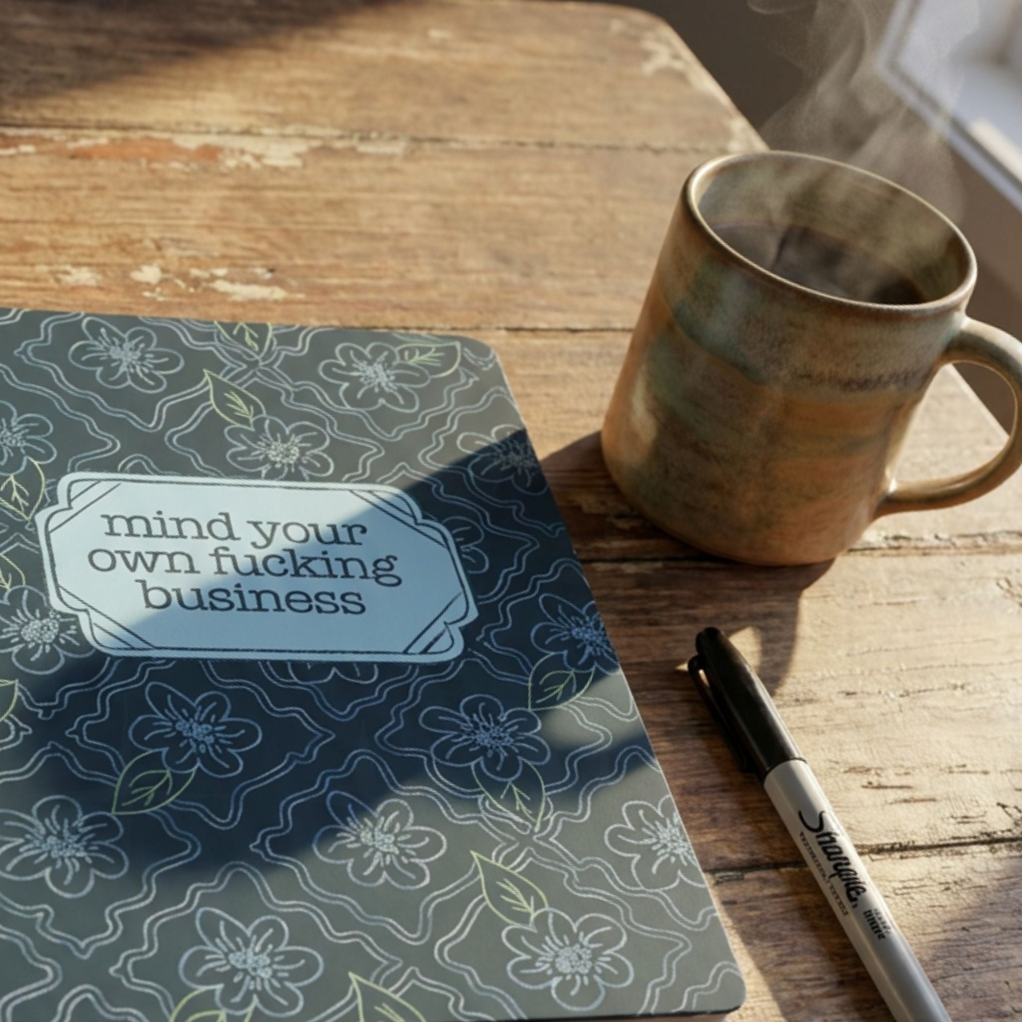 Profanity floral journal on rustic wooden table beside a steaming ceramic mug and black Sharpie marker in warm natural lighting