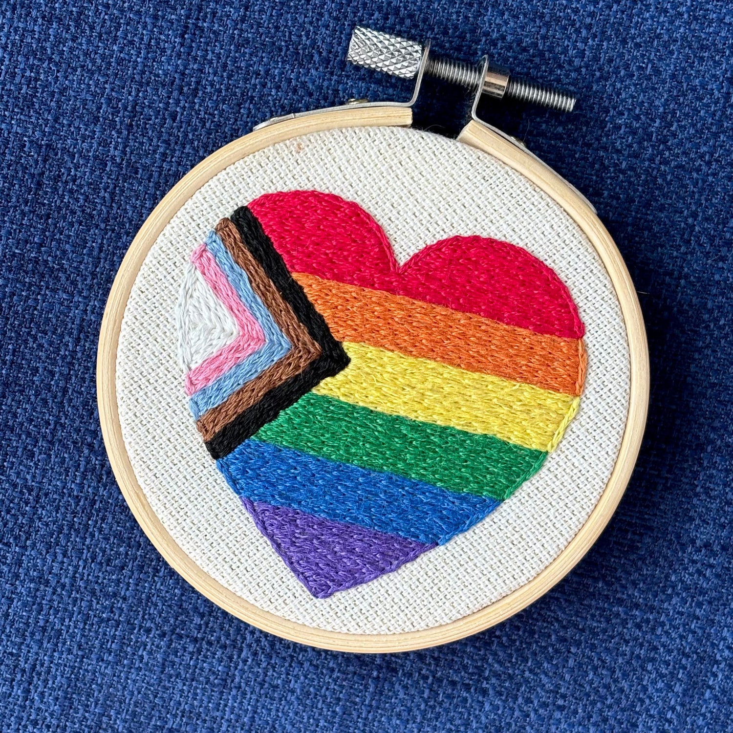 Heart-shaped progress pride flag hand embroidered onto cream colored linen. Displayed in a light wooden embroidery hoop as wall art or a christmas ornament.