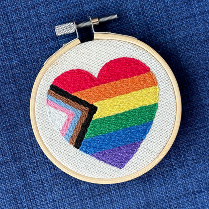 Heart-shaped progress pride flag hand embroidered onto cream colored linen. Displayed in a light wooden embroidery hoop as wall art or a christmas ornament.