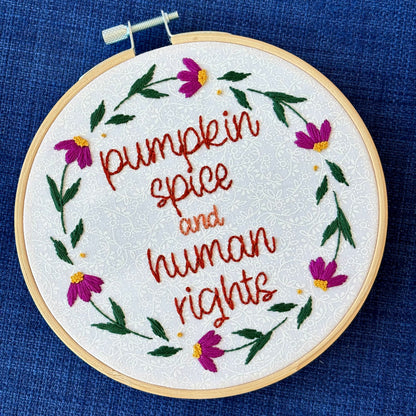 "Pumpkin spice and human rights". Handwriting font text hand embroidered in two shades of medium orange. Surrounded by a floral wreath of bright pink flowers with yellow centers and green leaves and stems. Framed in light wood embroidery hoop and displayed as wall art.