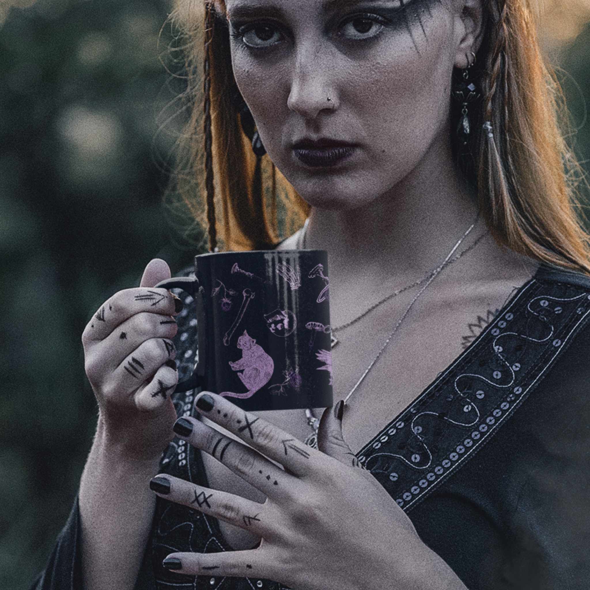 Woman with dark lipstick and tattooed hands holding a purple witchcore mug outdoors at dusk