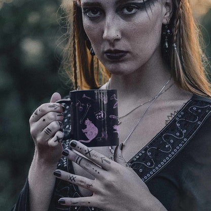Woman with dark lipstick and tattooed hands holding a purple witchcore mug outdoors at dusk