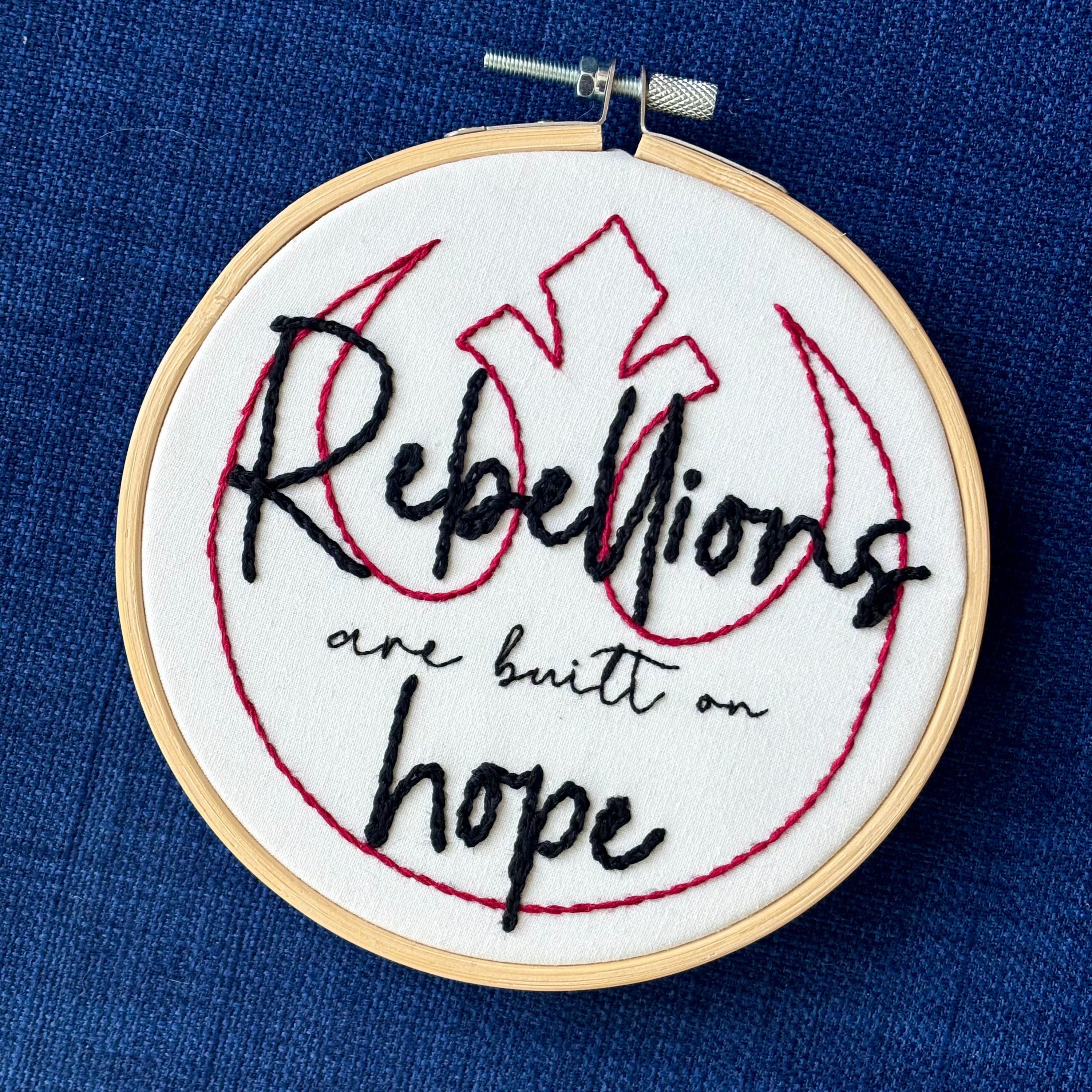 "Rebellions are built on hope." Text is in two handwriting fonts and hand embroidered in black embroidery thread. Words are centered on top of a red outline of the Star Wars Rebel Alliance symbol. Fabric is white cotton. Framed in light wood embroidery hoop.