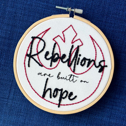 "Rebellions are built on hope." Text is in two handwriting fonts and hand embroidered in black embroidery thread. Words are centered on top of a red outline of the Star Wars Rebel Alliance symbol. Fabric is white cotton. Framed in light wood embroidery hoop.