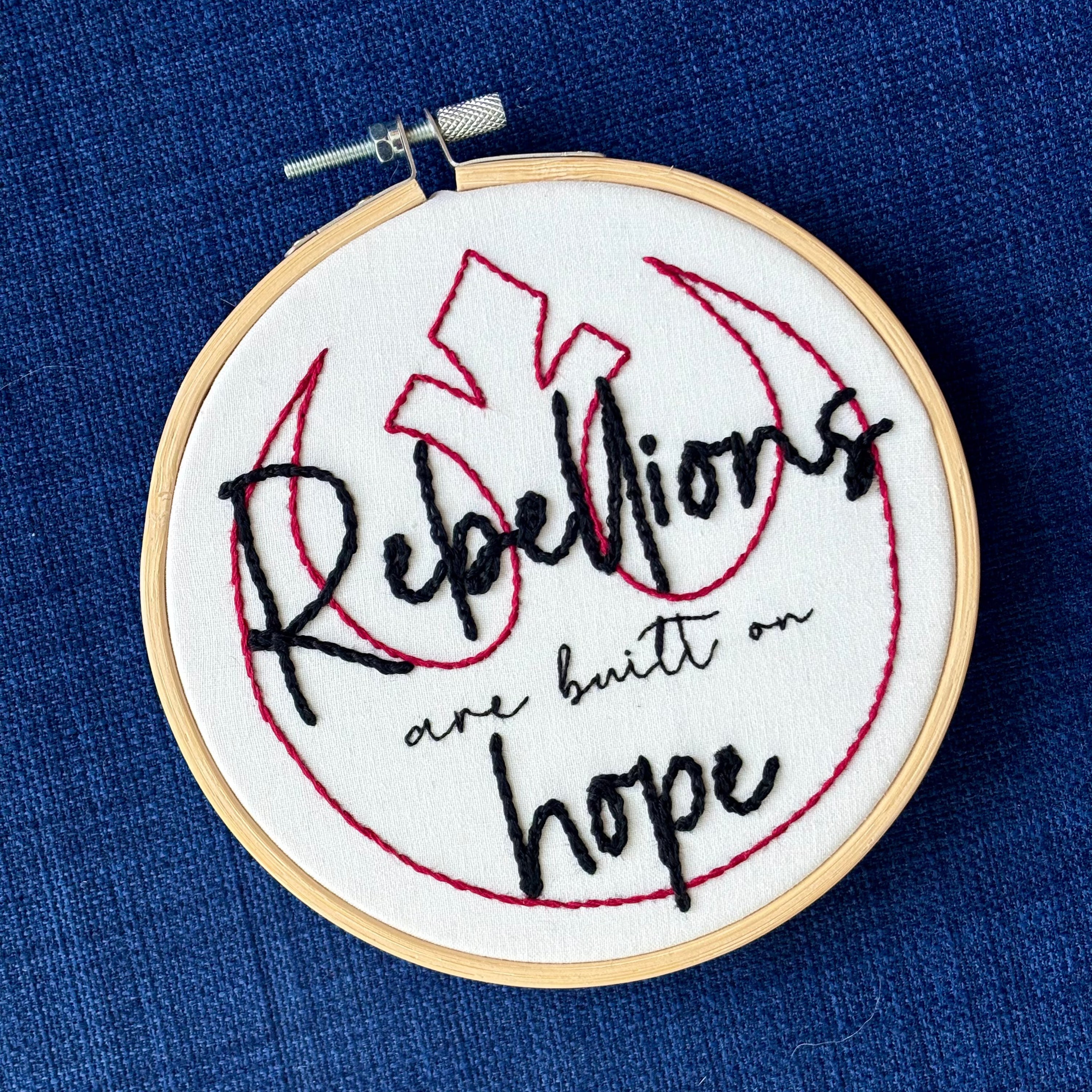 "Rebellions are built on hope." Text is in two handwriting fonts and hand embroidered in black embroidery thread. Words are centered on top of a red outline of the Star Wars Rebel Alliance symbol. Fabric is white cotton. Framed in light wood embroidery hoop.