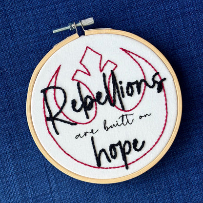 "Rebellions are built on hope." Text is in two handwriting fonts and hand embroidered in black embroidery thread. Words are centered on top of a red outline of the Star Wars Rebel Alliance symbol. Fabric is white cotton. Framed in light wood embroidery hoop.