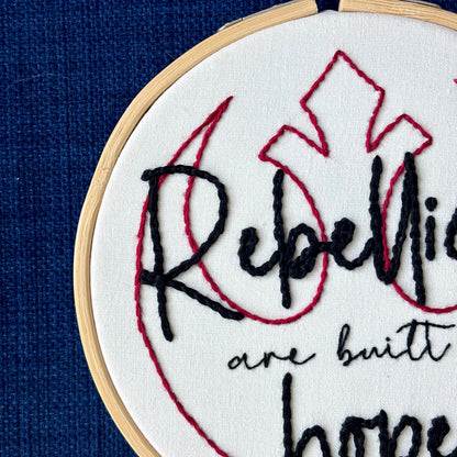 "Rebellions are built on hope." Text is in two handwriting fonts and hand embroidered in black embroidery thread. Words are centered on top of a red outline of the Star Wars Rebel Alliance symbol. Fabric is white cotton. Framed in light wood embroidery hoop.