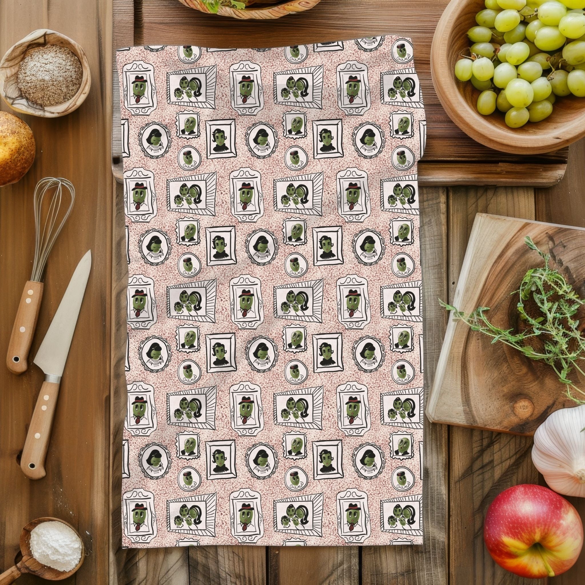 Dusty rose illustrated olive tea towel styled flat on rustic wood surface with kitchen props including whisk, knife, grapes, and apple