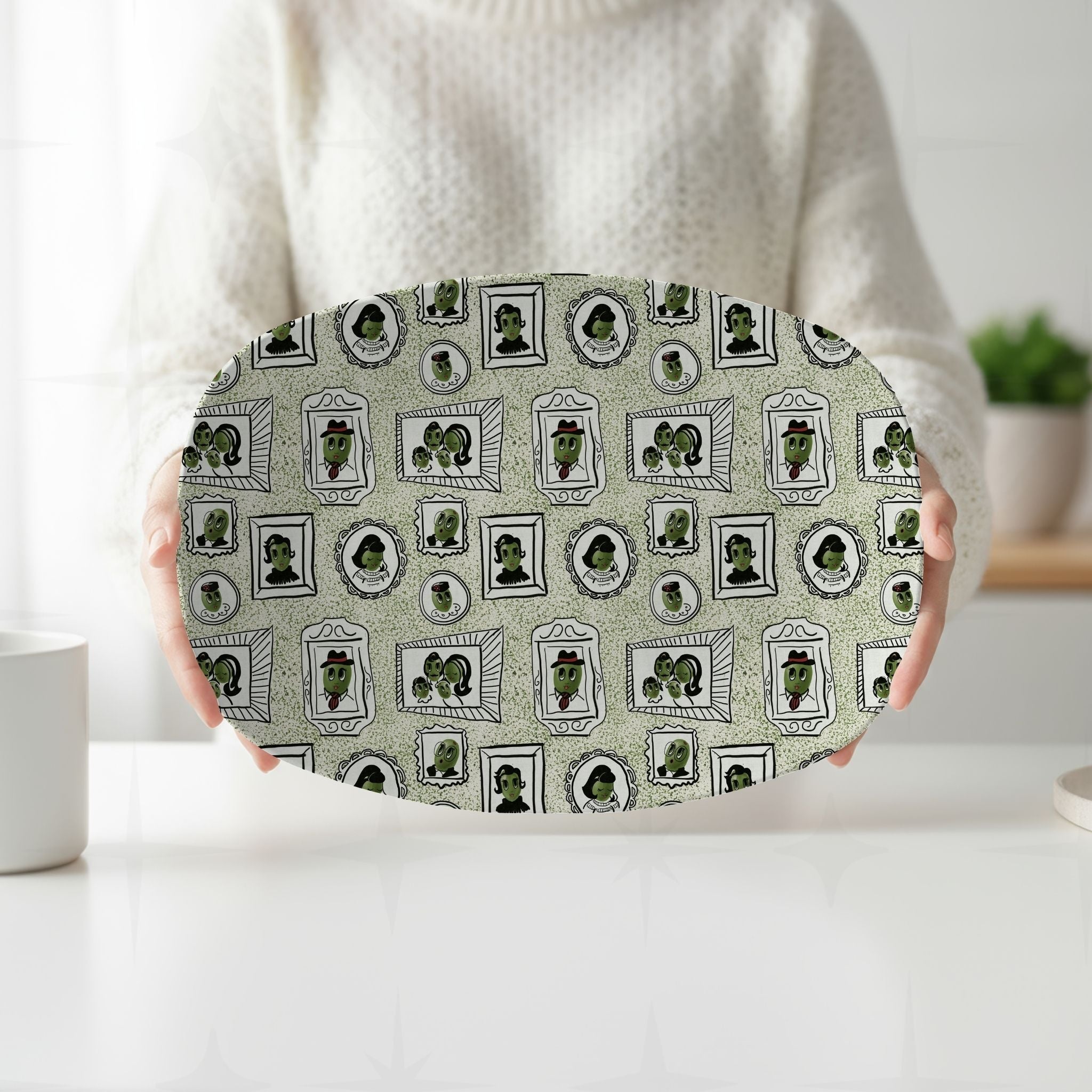 Person in cream sweater holding retro olive portrait serving platter in green colorway with both hands at white kitchen counter, full mid-century gallery wall pattern visible