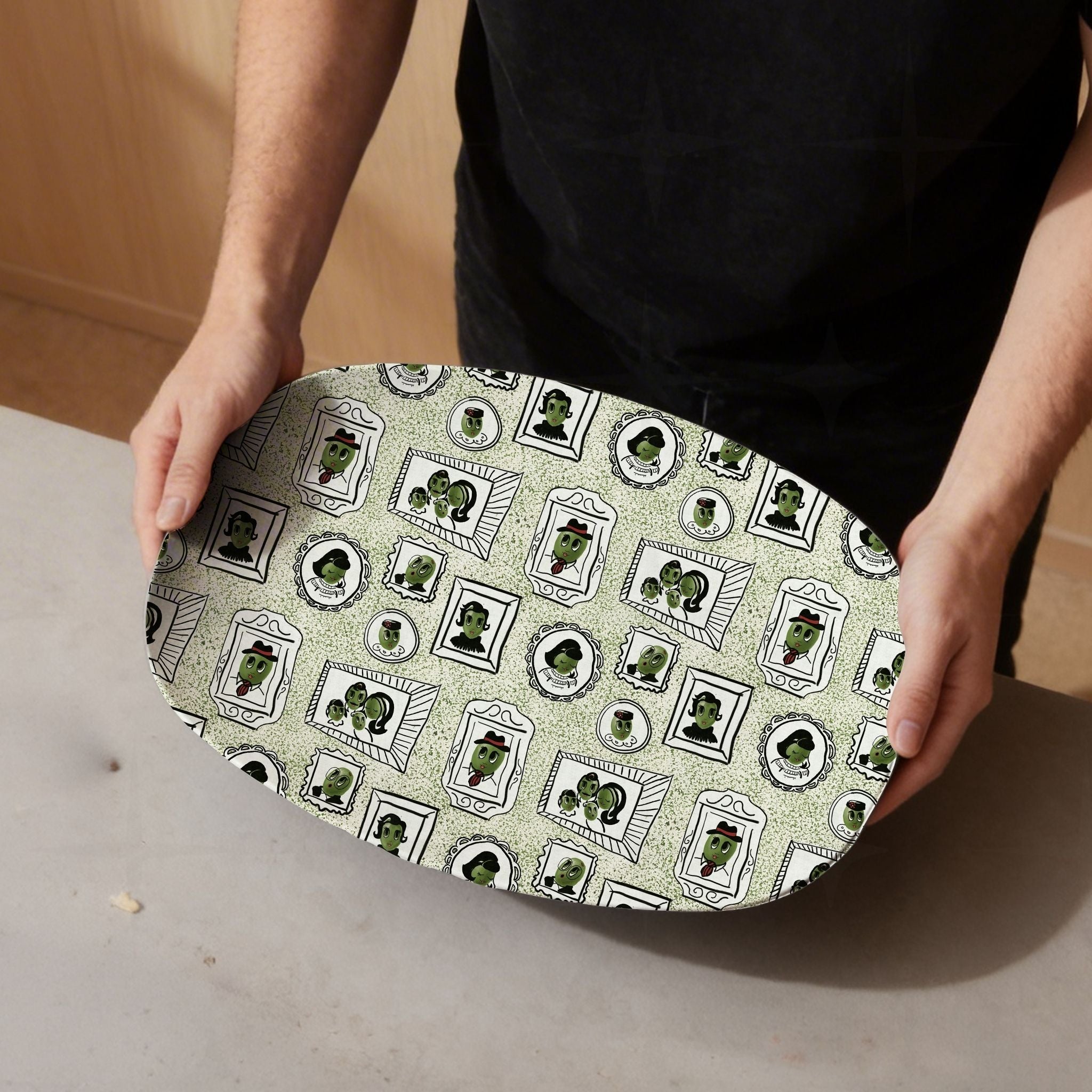 Man in black t-shirt holding retro olive portrait serving platter in sage green colorway tilted toward camera showing full mid-century gallery wall framed olive pattern