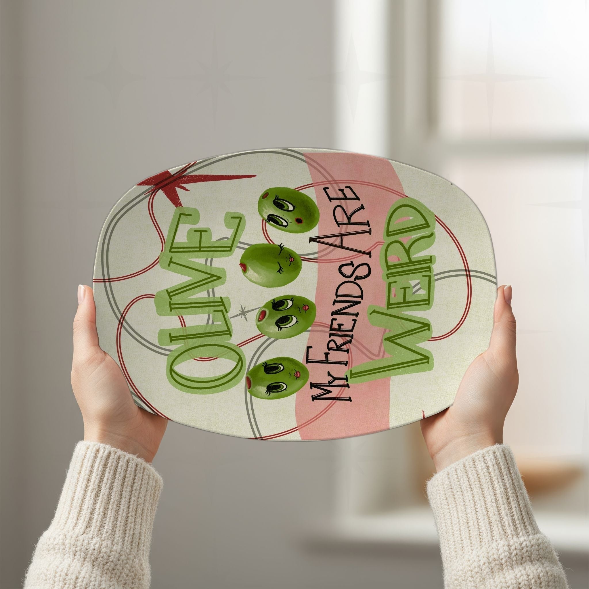 Two hands holding retro olive pun platter overhead against bright white background, showing full "Olive my friends are weird" mid-century illustrated design with atomic starburst motif and oval line framing