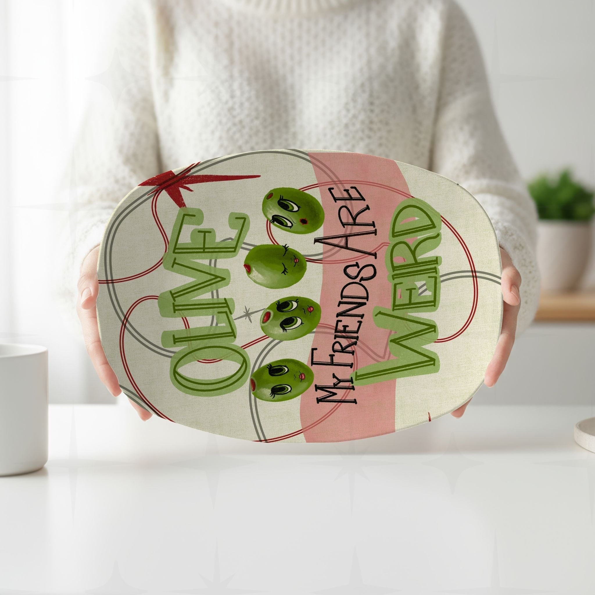 Person holding retro mid-century olive pun serving platter with both hands in front of white kitchen background, full illustrated design visible with blush pink color block and sage green dimensional lettering