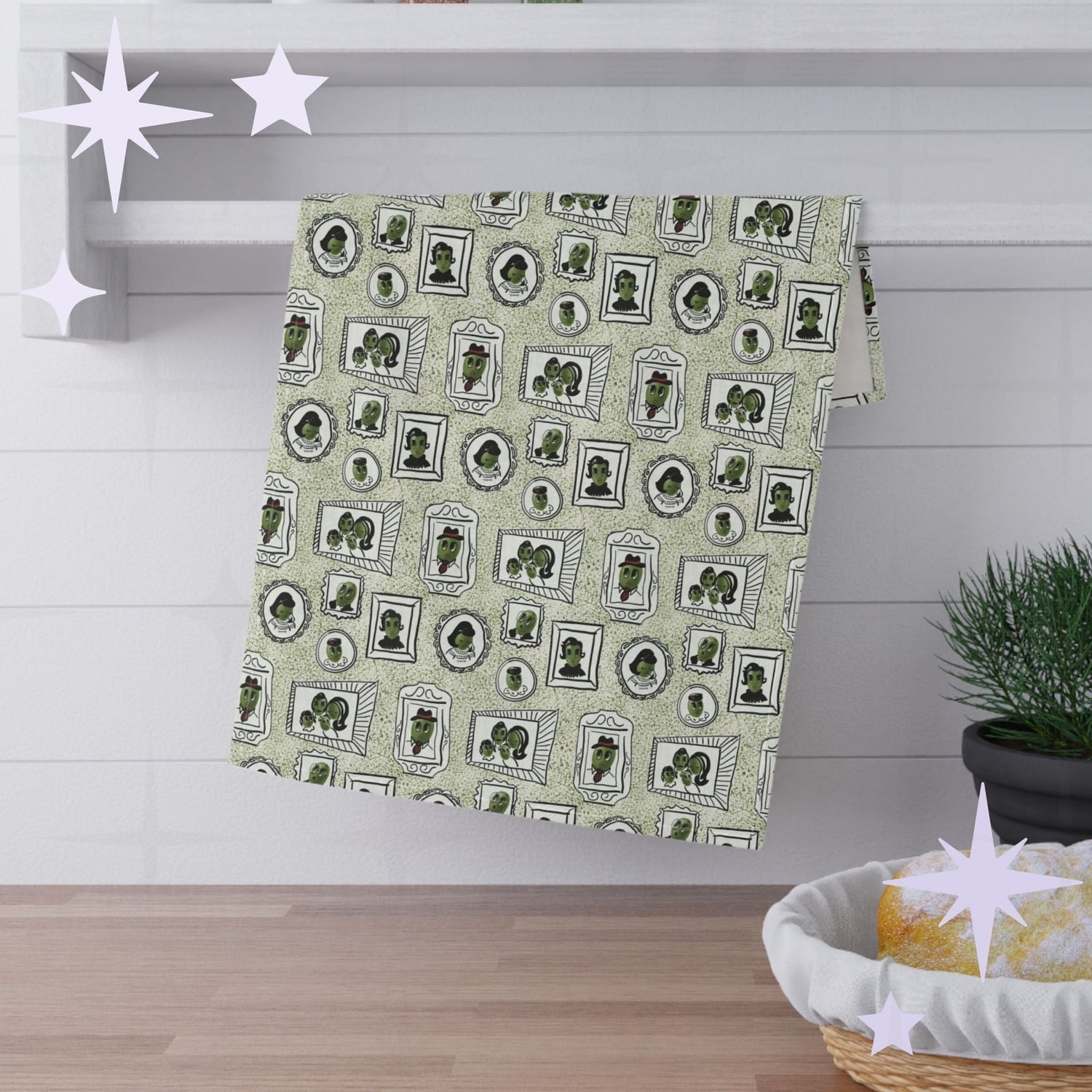 Sage green illustrated olive tea towel hanging folded over white kitchen shelf, mid-century portrait pattern visible, styled with pine sprig and bread basket