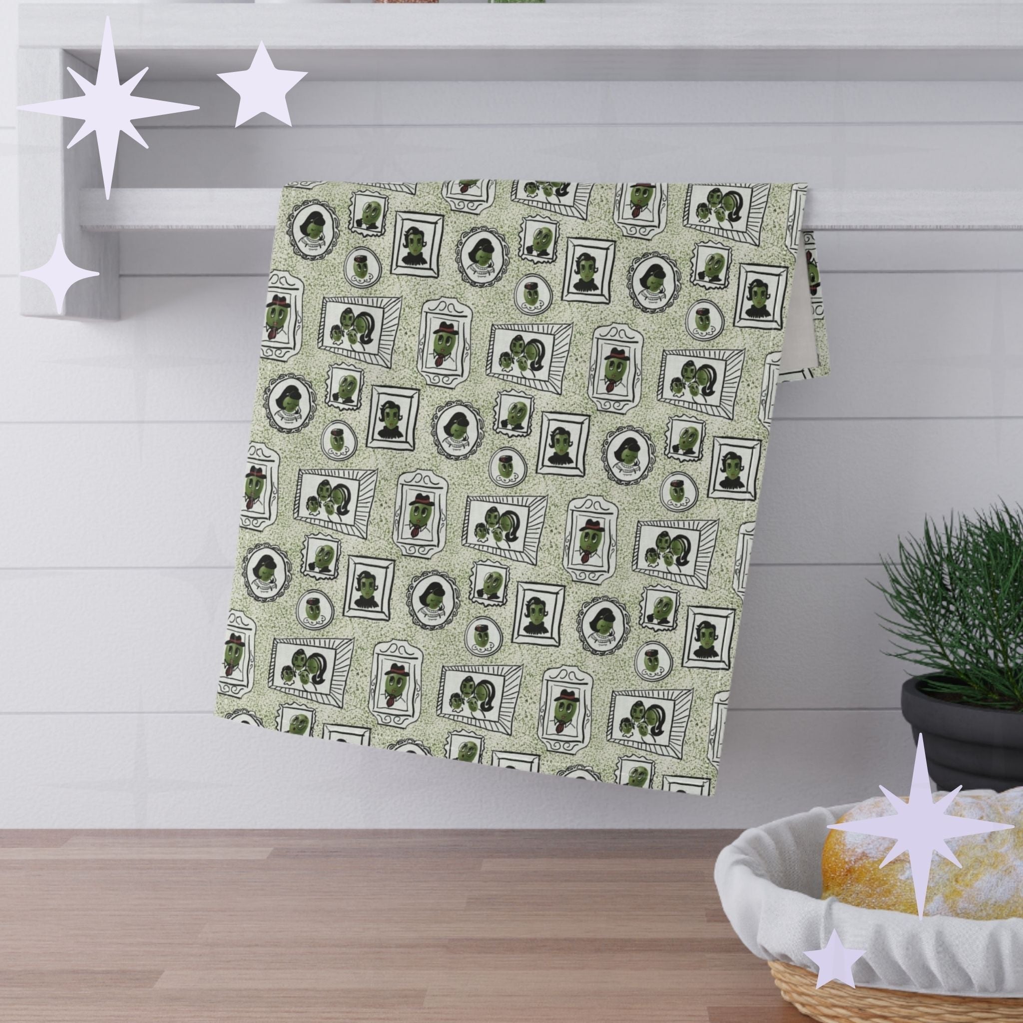 Sage green illustrated olive tea towel hanging folded over white kitchen shelf, mid-century portrait pattern visible, styled with pine sprig and bread basket