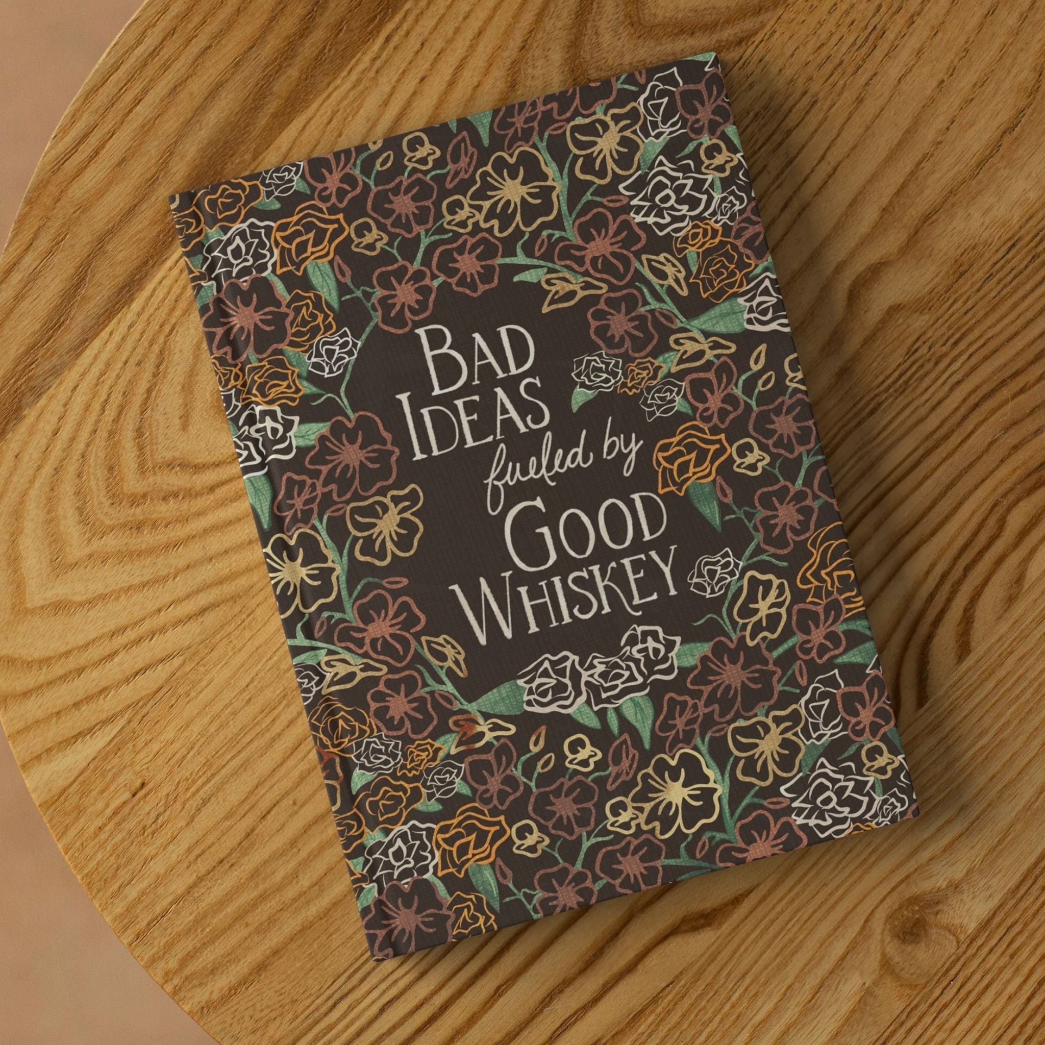 Maximalist floral whiskey journal lying on round wooden surface showing front cover with dark wildflower pattern and vintage typography