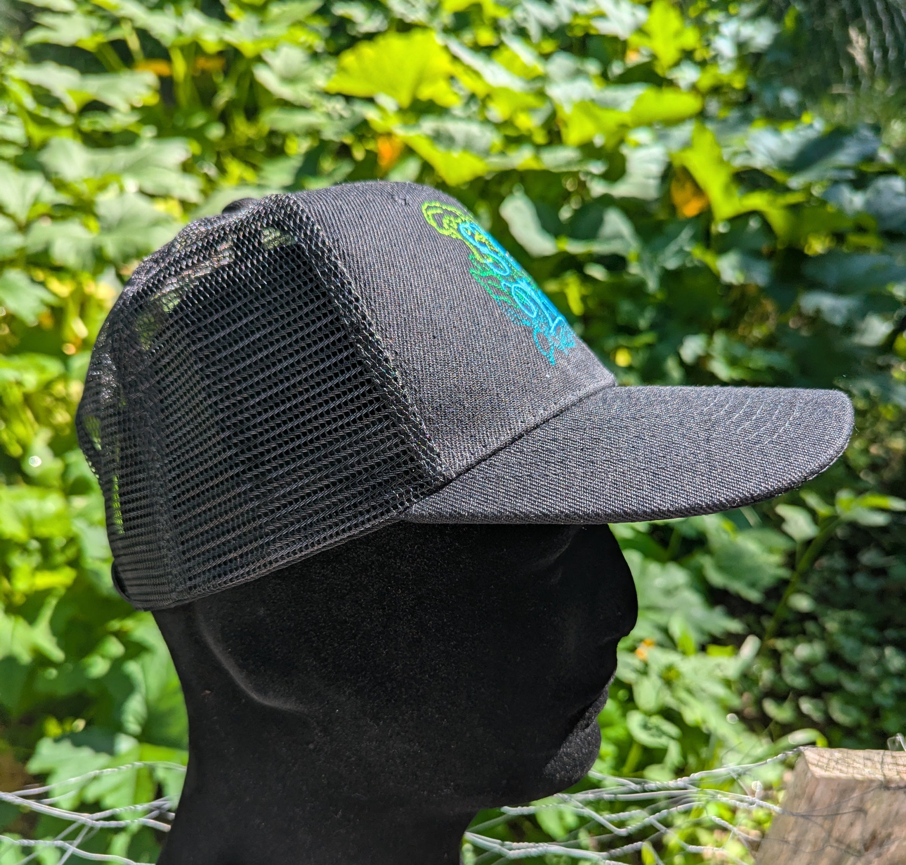A black trucker-style cap with a black mesh back and a small green and blue graphic on the front panel, shown on a black mannequin head outdoors with a garden background.