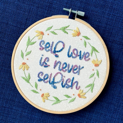 "self love is never selfish." Quote hand embroidered in color changing thread of pink, lavender, light blue, and royal blue. Surrounding the quote is a floral wreath of yellow and orange daisies. Sewn on white-on-white cotton. Finished wall art is framed in a light wood embroidery hoop.